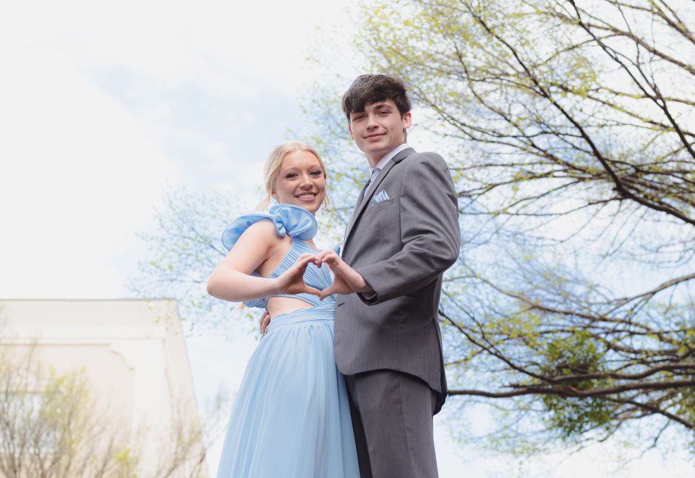 A couple in formal attire poses outdoors together in a light blue dress and grey suit on a sunny spring day.