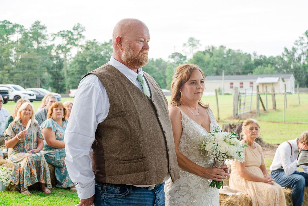 A couple in rustic wedding attire stands together during their outdoor country ceremony with guests seated on hay bales.