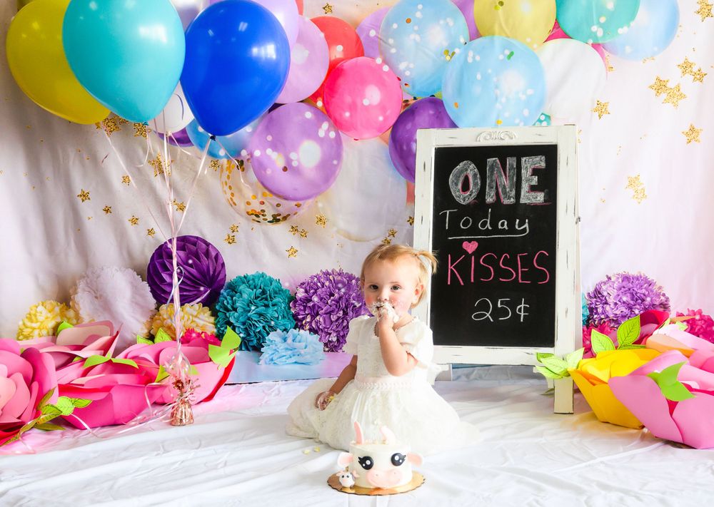 Colorful first birthday cake smash setup with balloons, chalkboard sign and party decorations against white backdrop.