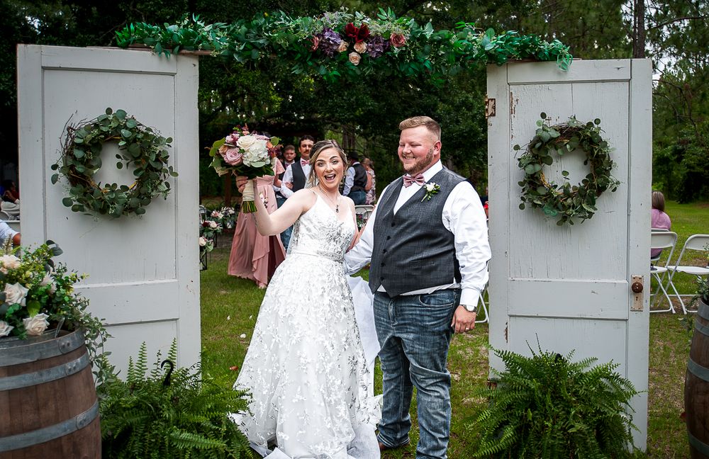 A joyful couple celebrates at their outdoor wedding ceremony between two white doors decorated with floral wreaths.