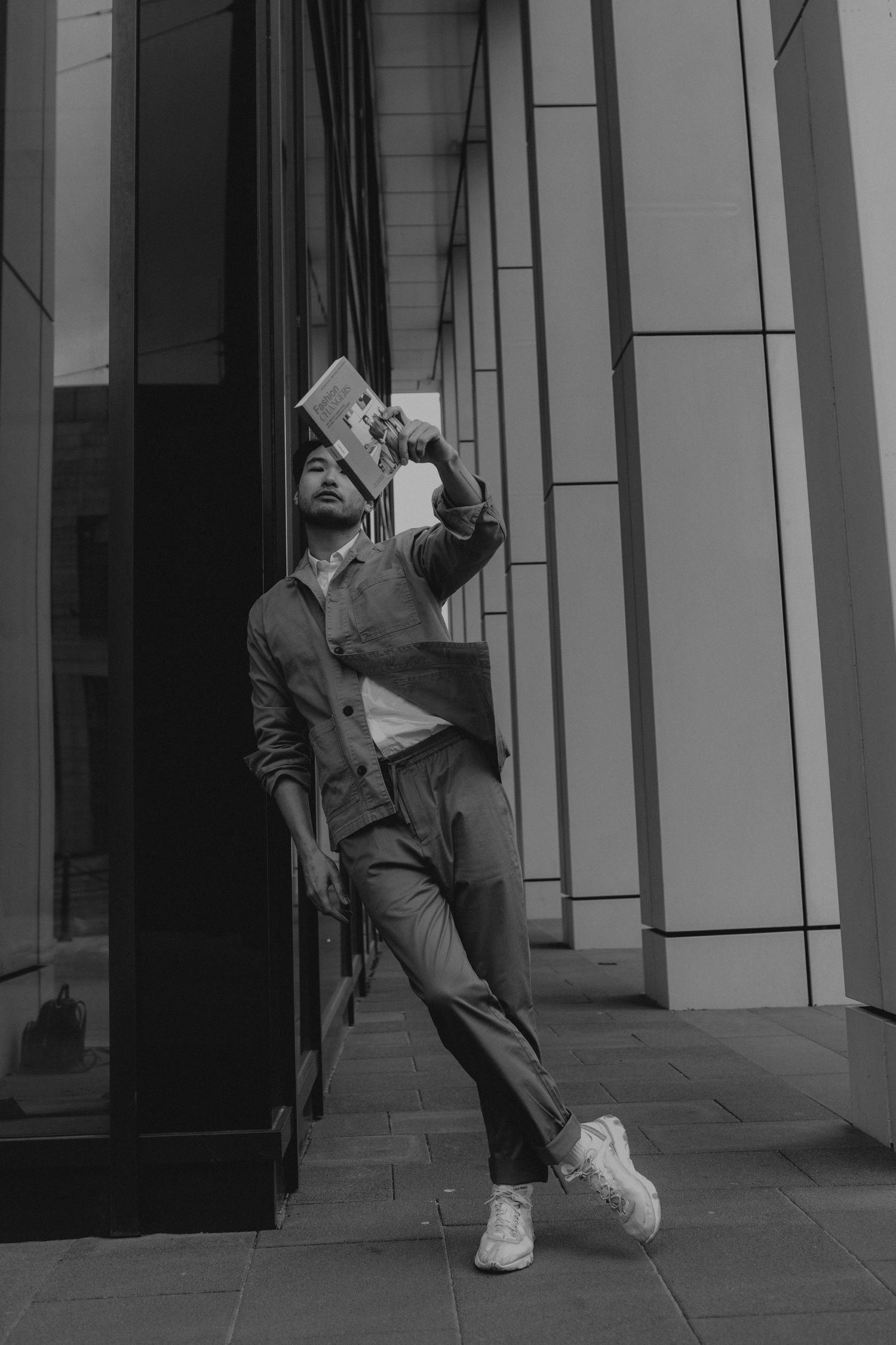 Artistic black and white photo of person in casual attire posing against modern building exterior.