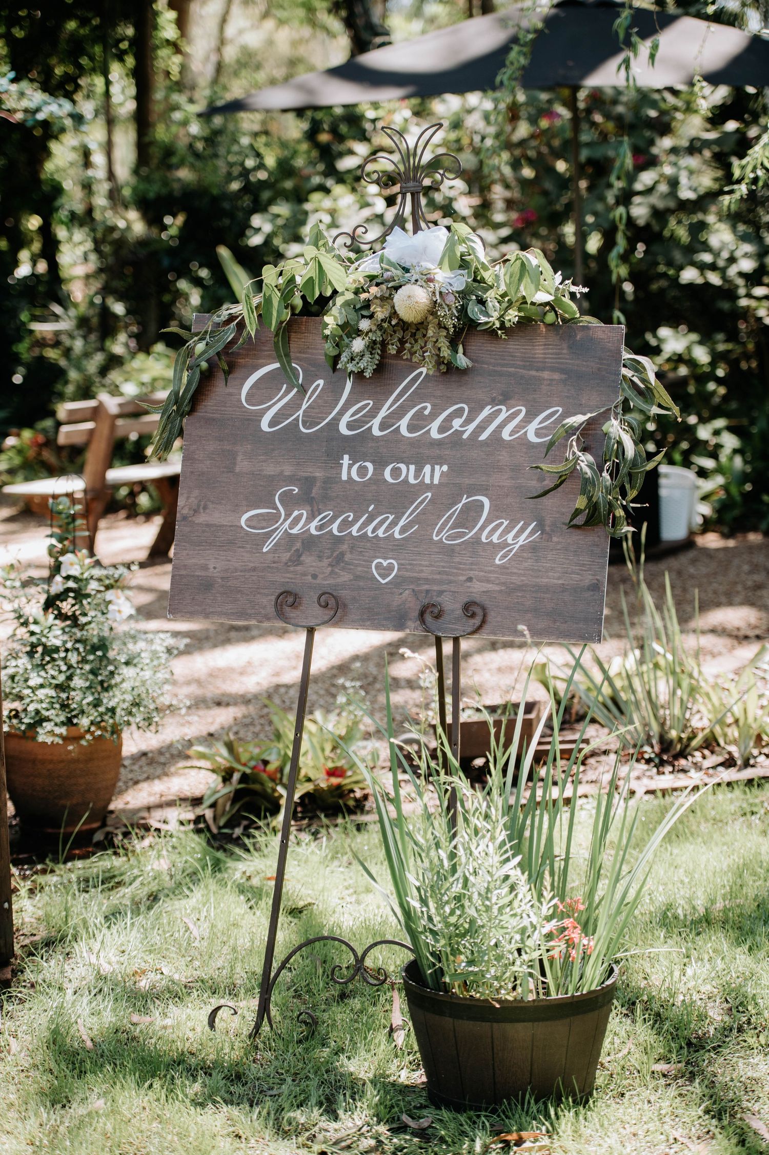 Rustic wooden wedding welcome sign decorated with greenery and florals stands in a garden setting.