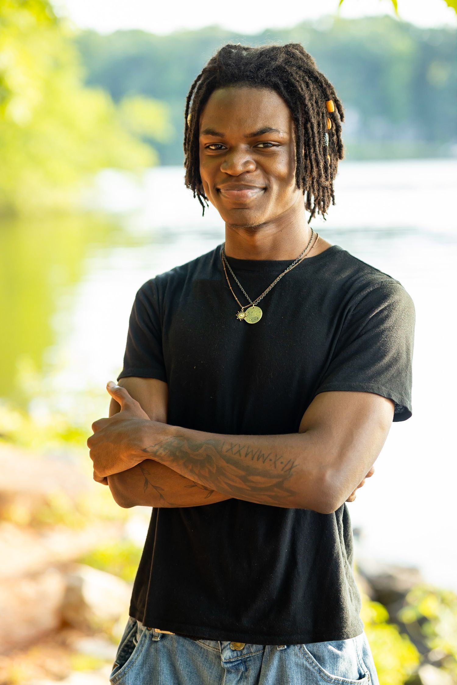 Person in black t-shirt with dreadlocks smiling outdoors by the water.