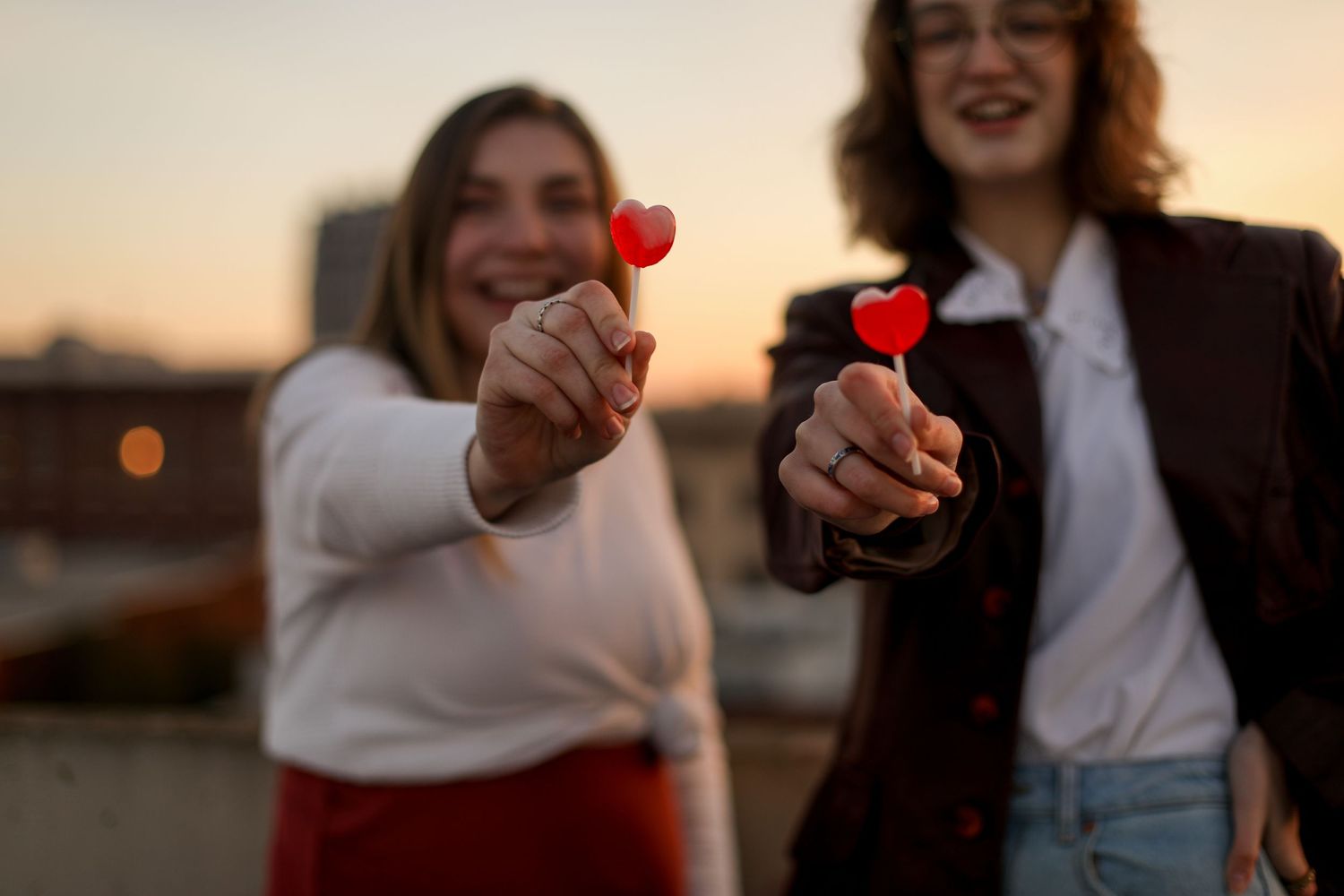 Two people holding red heart lollipops toward camera at sunset.