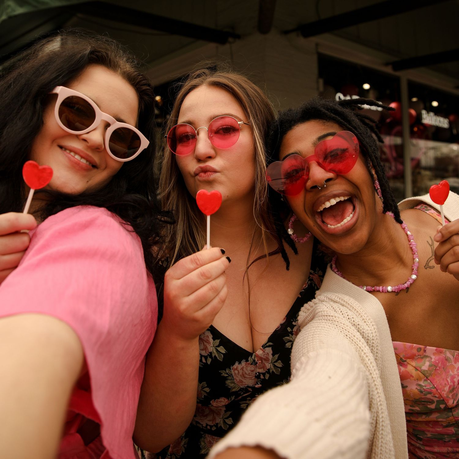 Three people posing with heart-shaped lollipops and pink sunglasses in a fun selfie.