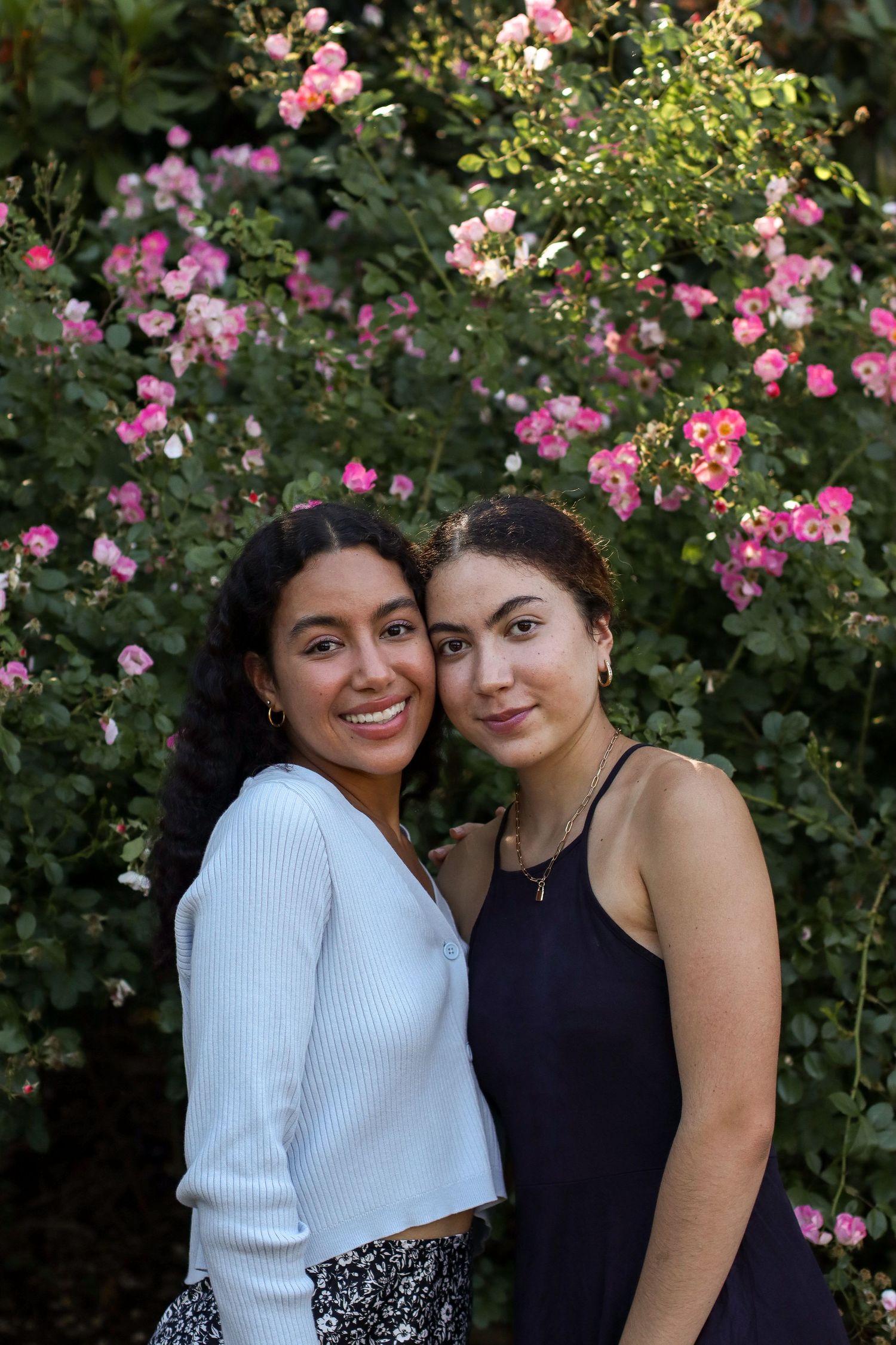 Two friends posing together in front of pink flowering rose bushes.