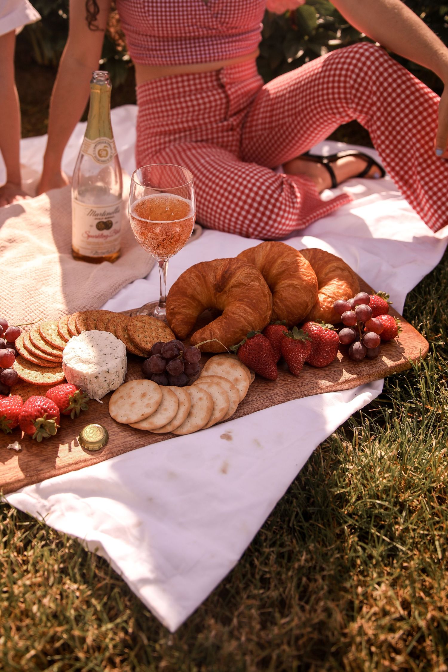 Picnic spread with croissants, fruit, and wine on a white blanket in dappled sunlight.