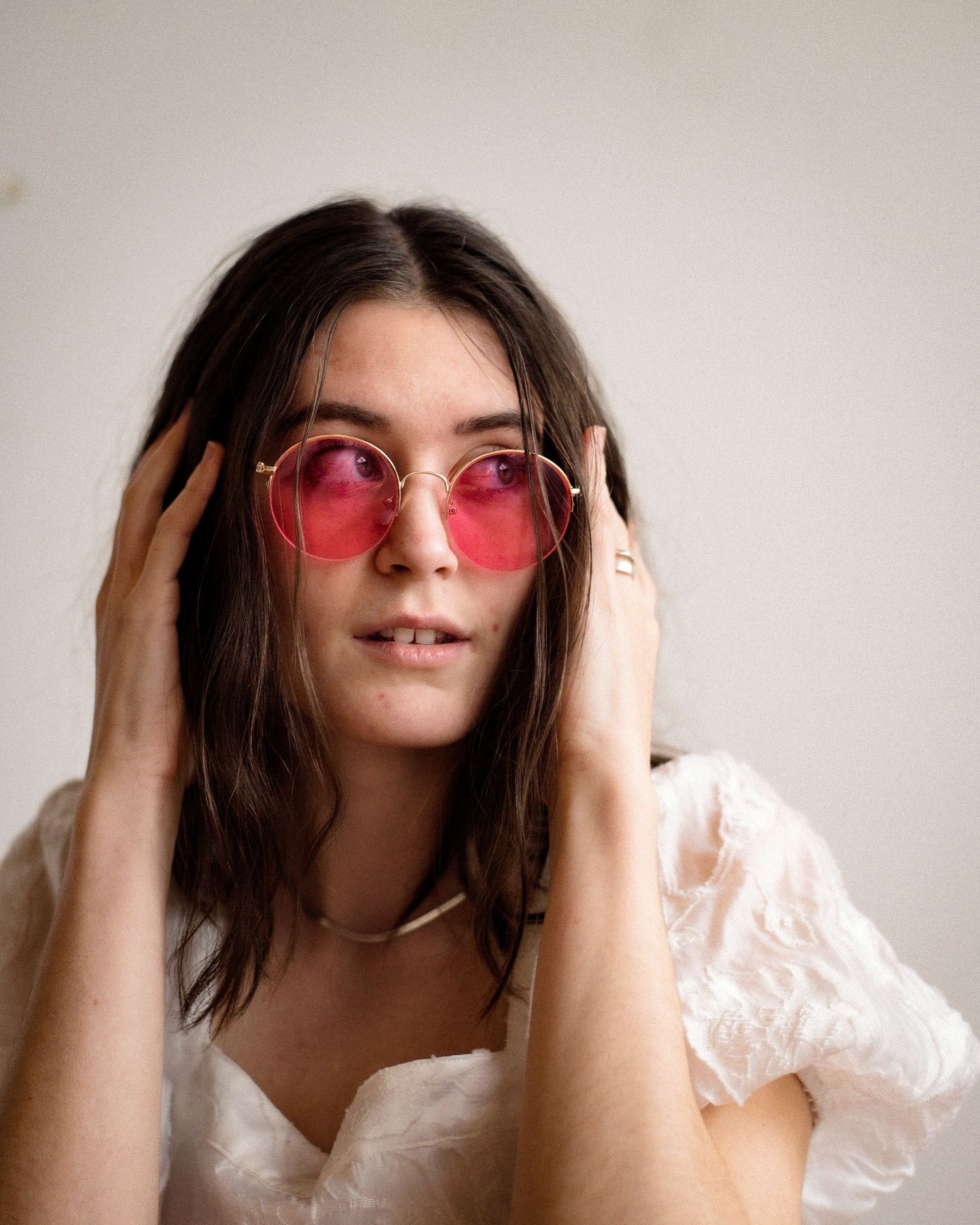 Portrait of person wearing pink tinted glasses and white lace top against light background.