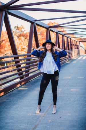 Stylish fall outfit with blue denim jacket, white top and black pants on metal bridge during sunset.