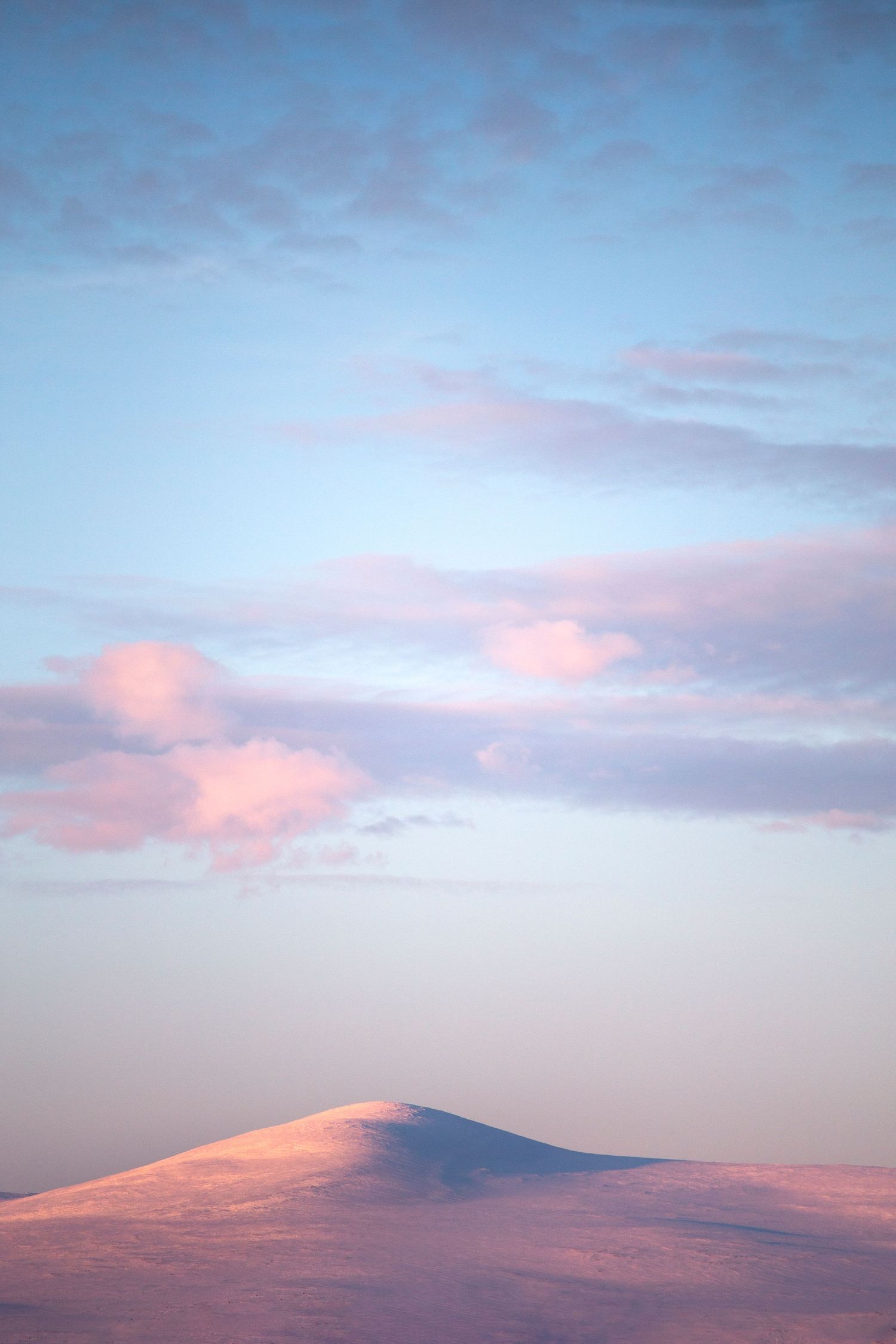 Snow-covered mountain peak bathed in soft pink light beneath a pastel blue sky with fluffy clouds.