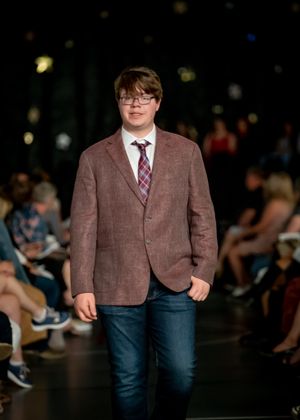 A model walks the runway in a brown blazer, plaid tie and jeans during a fashion show.