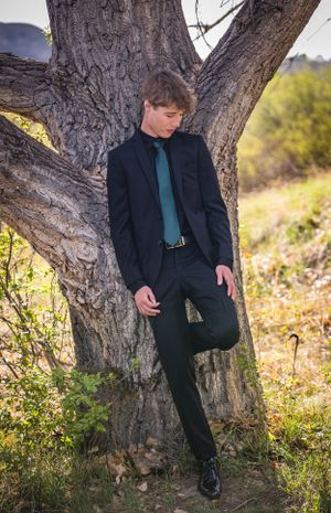 Formal portrait of person in dark suit and teal tie leaning against large tree trunk.