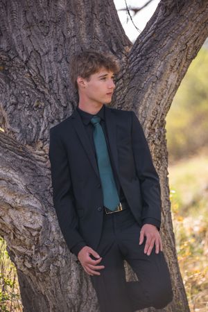 Portrait of person in navy suit with teal tie posing by textured tree trunk.