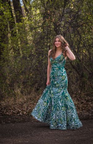 Person in flowing teal patterned gown poses on outdoor path surrounded by trees.