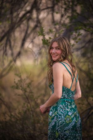 Person in strappy teal patterned dress smiles over shoulder in natural setting.