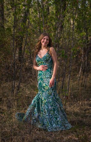 Person in fitted teal gown poses gracefully among trees in outdoor setting.