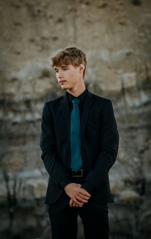 Young man in dark suit and teal tie poses against textured stone wall backdrop.