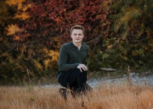 Person in dark green sweater kneeling in autumn field with colorful fall foliage.