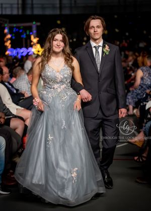 A couple walks the runway in formal wear - her in a gray gown, him in black suit.