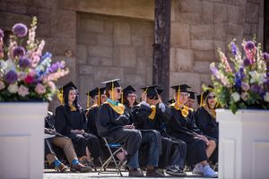 Graduates in black caps and gowns seated at commencement with floral arrangements.