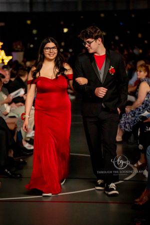 A couple walks down a runway in formal attire, with the woman wearing a stunning red gown and the man in a black suit.