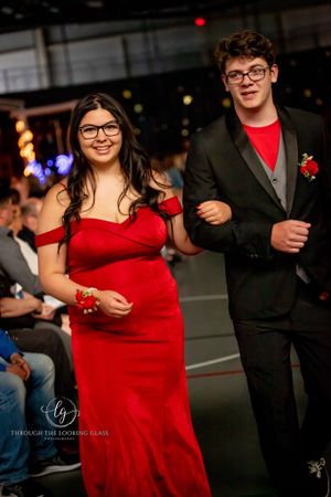 Elegant couple smiles while walking a fashion show runway, woman in red off-shoulder gown and man in dark suit.
