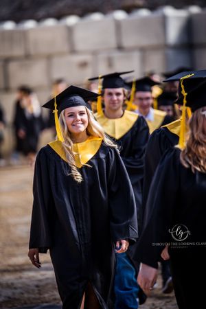Graduate in black cap and gown with yellow stole stands among fellow graduates during commencement ceremony.