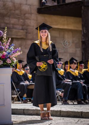 Graduate in black cap and gown with yellow stole poses by floral arrangement during graduation ceremony.