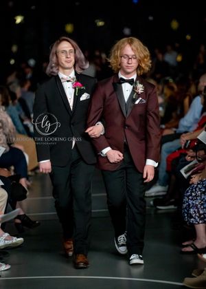 Two people walk runway wearing formal attire, one in black suit and one in burgundy suit with bow ties.