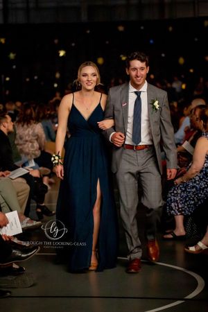 Couple walks fashion runway, woman wearing navy gown with slit and man in gray suit with tie.