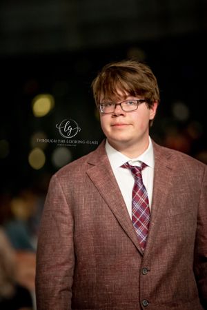 A person wearing glasses and a brown suit jacket with plaid tie poses for a formal portrait.