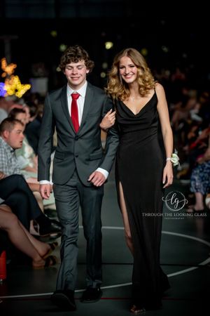 A couple walks arm in arm on a runway, with him in a dark suit and her in an elegant black gown.