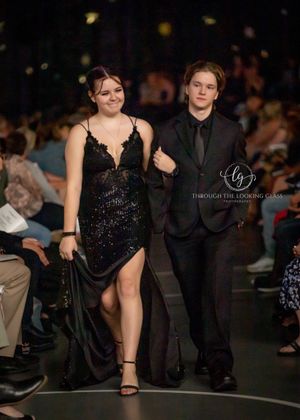 A couple walks the runway together, with her wearing a sparkly black gown and him in all black attire.