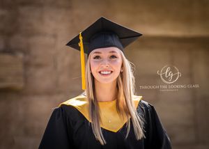 A graduate in traditional cap and gown smiles brightly against a stone wall background.