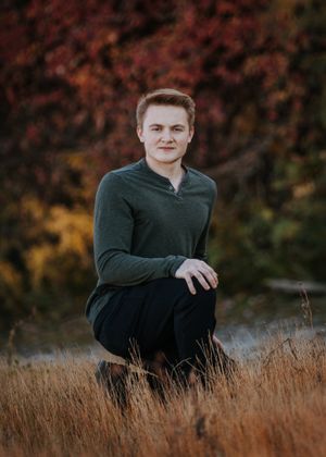 Portrait of person in green sweater kneeling in tall grass with autumn colors in background.
