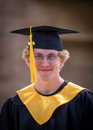 Graduate in traditional academic dress with black cap and yellow stole smiling at camera.