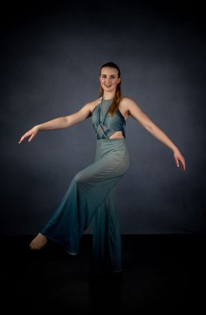 A dancer in teal performance attire strikes an elegant pose against a dark studio backdrop.