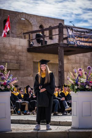 Graduate in black cap and gown standing between floral arrangements at outdoor graduation ceremony.