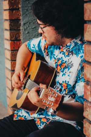 Musician in floral Hawaiian shirt playing acoustic guitar against brick wall background.