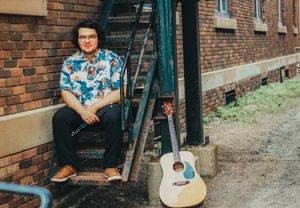 Guitarist in blue floral shirt sitting on metal stairs with acoustic guitar.