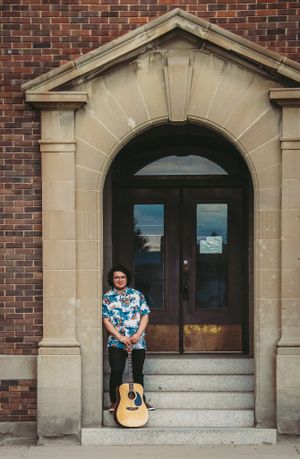 Musician with acoustic guitar standing in arched doorway of brick building.