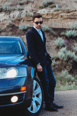 Person in dark suit and sunglasses poses beside blue car near rocky desert landscape.