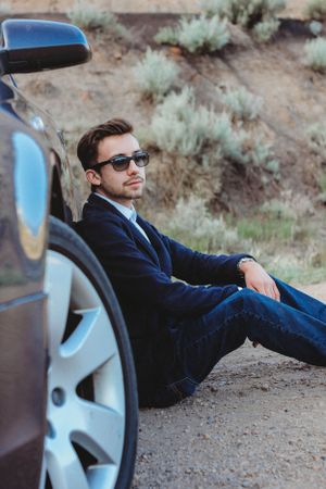 Person in navy suit and sunglasses sits against blue car door in desert setting.