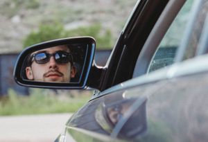 Reflection of driver wearing sunglasses captured in car side mirror.