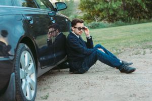 Person in dark suit sitting on ground beside parked car.