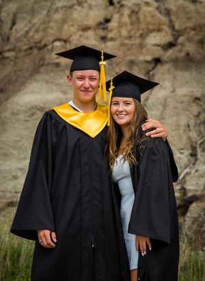 Two graduates in black and gold regalia pose together in front of a rocky cliff.