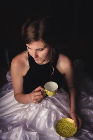 A woman in black dress sits on white fabric with a vintage yellow teacup.