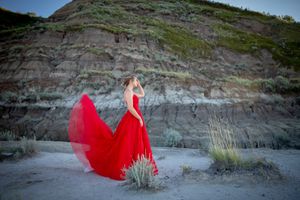 Red dress catches wind against dramatic desert cliff backdrop at dusk.