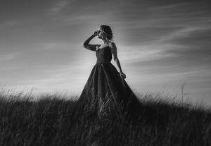 Dramatic black and white silhouette of a figure in an elegant ballgown standing in a field against a cloudy sky.