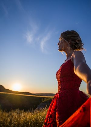 Silhouetted figure in red dress stands against sunset sky in rural landscape.